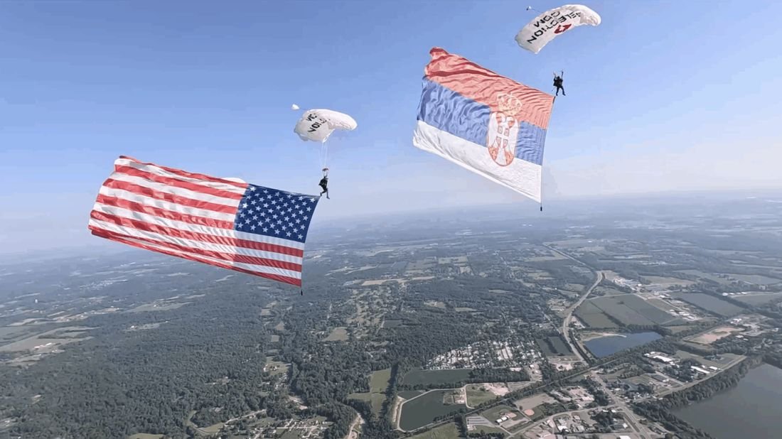 American and Serbian flags flying under parachutes over the countryside